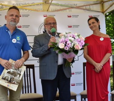 trzy osoby stoją na scenie, mężczyzna po lewej stronie trzyma teczkę, mężczyzna po środku trzyma kwitay przemawia do...