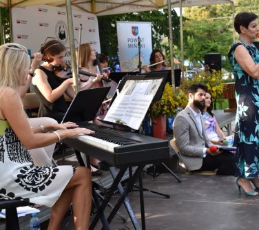 zespól siedi nakrzesłach na zadaszonej scenie, grają na instrumentach, kobieta na przodzie śpiewa, obok na krzesłach siedzi...