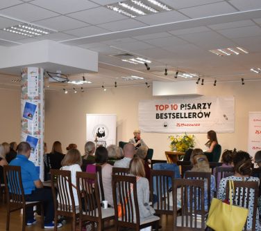 słuchacze siedzą na krzesłach przed nimi mini scena na któej siedza dwie kobiety, po lewej stronie kobieta przemawia do...