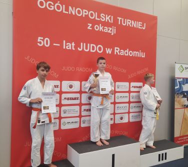 trzech chłopców pozuje do zdjęcia stojąc na podium, w rękach trzymają dyplomy, w tle baner ogólnopolski turniej z okazji...