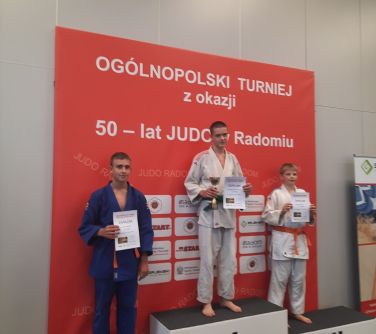 trzech nastolatków pozuje do zdjęcia stojąc na podium, na pierwszym miejscu trzyma puchar i dyplomw dłoniach, za drugie i...