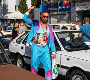 mężczyzna w kolorowym dresie z reklamówka w jednej dłoni, druga dłoń podniesiona do góry trzyma magnetofon przy uchu,...