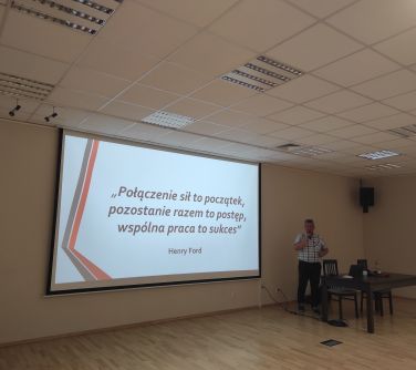 na ścianie wisi ekran na którym wyświetlany jest tekst, po prawej stronie przed stołem i obok krzesła stoi mężczyzna i...