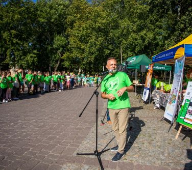 mężczyzna przemawia na dziedzińcu miejskiego domu kultury za nim namioty z banerami i stołami z gadżetami, patrzy w tył,...