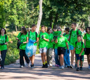 grupa dzieci w zielonych koszulkach maszeruje alejką w parku, mają założone foliowe rękawiczki, niektórzy trzymają worki na...