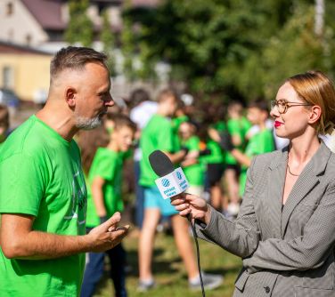 mężczyzna po lewej w zielonej koszulce udziela wywiadu kobiecie przemawiając do mikrofonu, w tle dzieci, drzewa i domy