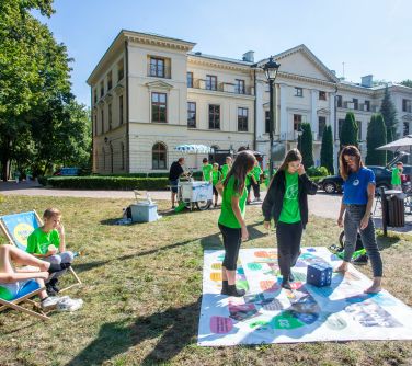plansza na trawie na planszy trzy osoby grają dużą kostką , po lewej stronie na dwóch leżakach siedzi dwóch chłopców, w tle...