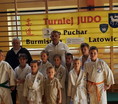 dzieci w strojach od judo oraz dwóch mężczyzn pozują do zdjęcia w tle drabinki na hali soportowej i zawieszony na nich...
