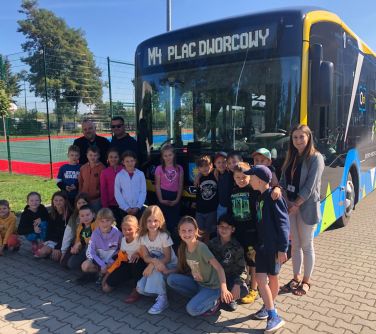 grupa osób pozuje na przodzie autobusu elektrycznego M4 Plac Dworcowy, po lewej stronie za siatką boisko i drzewa