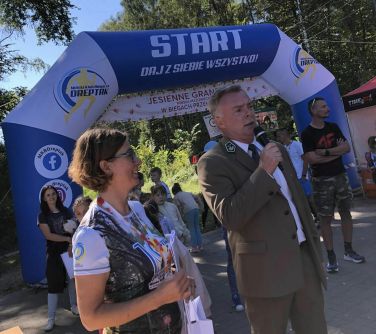 mężczyzna w garniturze z nadleśnictwa przemawia do mikrofonu, obok stoi usmiechnięta kobieta, w tle dmuchany duży balon...