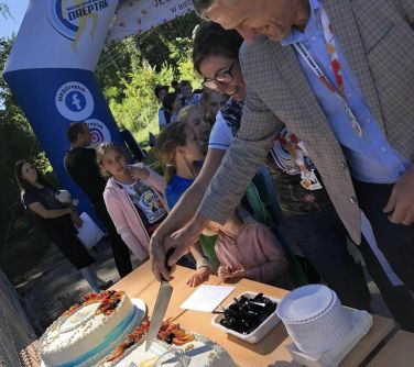 uśmiechnięta kobieta i uśmiechnięty mężczyzna kroją tort po lewej stronie dmuchany duży balon start obok dzieci