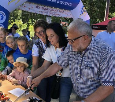 trzy osoby trzmaja nóż w rękach przygotowują się do krojenia tort, po lewej stronie duży dmuchany balon Start, obok dzieci,...