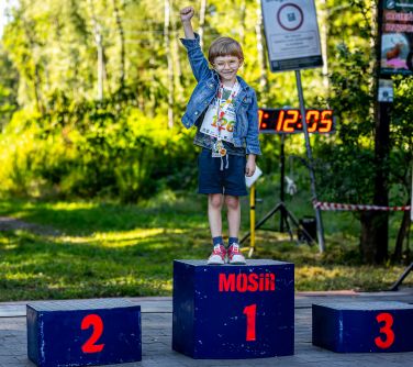 trzy oddzielne stopnie podium, na pierwszym miejscu stoi uśmiechnięte dziecko z medalem na szyi, trzyma prawą dłoń...