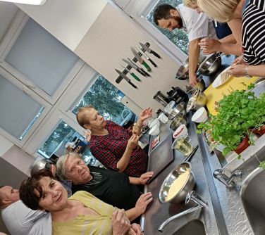 osoby w kuchni szkolnej przygotowują posiłki, przed nimi na stołach metalowych ze zlewem i kranem stoją w miskach i kubkach...