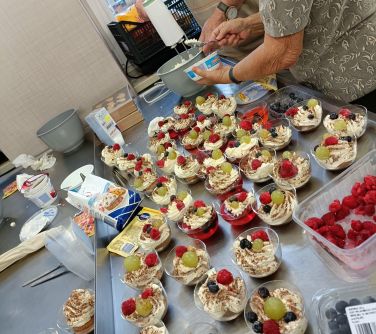 na stole w plastikowych małych pojemnikach stoją deserki z bitą śmietaną, galaretką i owocami, obok produkty, w tle dwie...