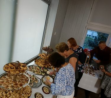 osoby częstują się poczęstunkiem ustawionym na stołach przykrytych białymi obrusmi. Jedzenie ułożone na metalowych tacah i...