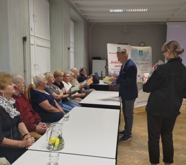 mężczyzna rozdaje laurki osobom które siedzą na krzesłach za stołami, w tle dwa roll-upy, po prawej stronie tyłem stoi...