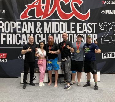 sześć osób (pięciu mężczyzn, jedna kobieta) poozują do zdjęcia stojąc przed podium, w tle baner ADCC powered by pitbull