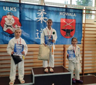 trzy dziewczynki stoją na podium trzymają dyplomy w dłoniach i siatki, w tle baner zawieszony na drabinkach w hali