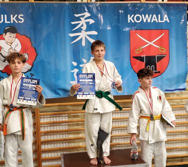 trzech chłopców stoi na podium obok dziewczynka, dwóch chłopców trzyma dyplomy, w tle baner zawieszony na drabinkach w hali...