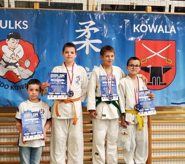 czterech chłopców stoi na podium trzymają dyplomy w dłoniach, za nimi w tle baner wisi na drabinkach na hali szkolnej, po...