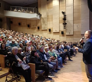 po prawej stronie stoi przed sceną i przemawia do mikrofonu przed publicznością