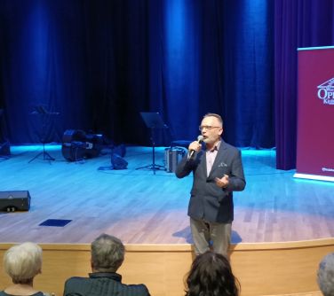 mężczyzna przemawia do mikrofonu stojąc przed sceną, w tle za nim na scenie instrumenty po prawej stronie roll-up...