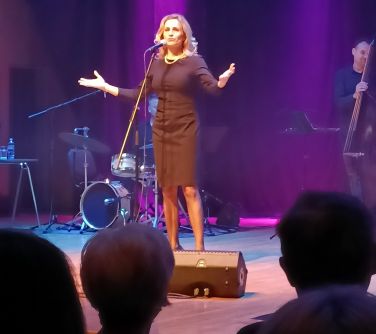 kobieta na scenie z rozłożonymi rękoma śpiewa do mikrofonu, w tle zespół