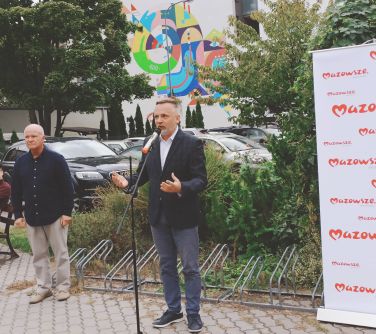 mężczyzna przemawia do mikrofonu, po lewej stronie roll-up mazowsza, po prawej drugi mężczyzna, za nimi stojaki dla rowerów