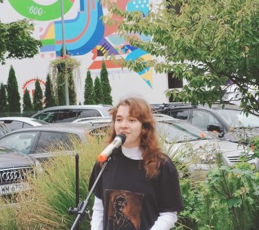 nastolatka przemawia do mikrofonu, w tle samochody i mural na ścianie budynku