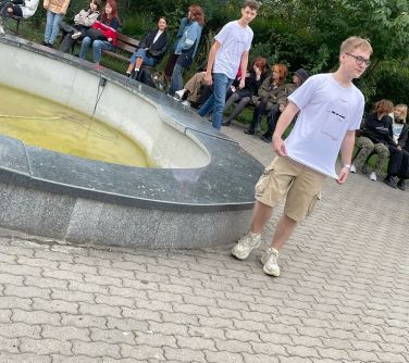 dwóch nastolatków przechadza się naokoło fontanny, w tle osoby siedza na ławkach