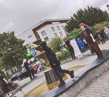 kilkoro nastolatków rpzadza się po krawędzi fontanny, w tle osoby, auta i budynki
