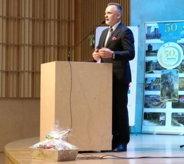 mężczyzna przemawia do mikrofonu stoją przed mównicą, za nim baner 50 lat gminy, przed mówincą pudelko łakoci...