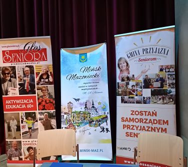 stoli przed nimi dwa krzesła, na stoliku dwie teczki i dwa długopisy obok nich, w tle trzy banery
