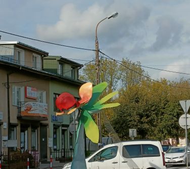 rzeźba kolorowy kwiat na rondzie, naokoło kwiaty, w tle biały samochód, po lewej stronie budynek i słup