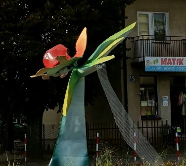 rzeźba kolorowego kwiatu naokoło posadzone kwiaty, w tle drzewo i budynek