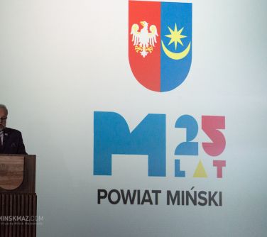 po lewej stronie mężczyzna przemawia do mikrofonu stojąc przy mównicy, w tle baner z herbem poniżej 25 lat Powiat Miński