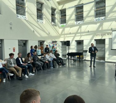aula po lewej stronie na krzesłach siedzą osoby, za nimi kilka osób stoi, na środku stoi i przemawia mężczyzna i stoi...