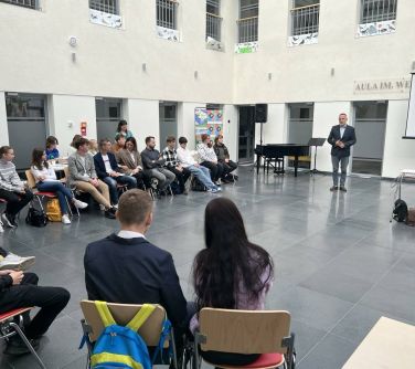 aula, naokoło na krzesłach siedza osoby, na przodzie stoi mężczyzna przemawia, obok niego fortepian