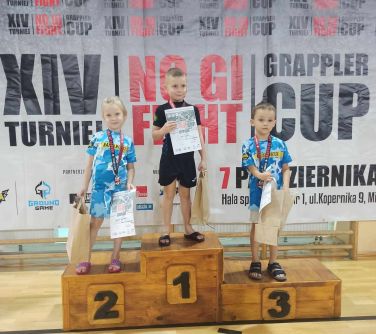 trójka dzieci pozuje a podium, trzymają w dłoniach dyplomy i torby papierowe, na szyjach medale, w tle baner VIV Turniej NO...
