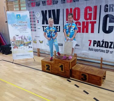 dwójka dzieci stoi na podium, na szyjach maja zawieszone medale, po lewej stronie roll-up Mińsk Mazowiecki, w tle XIV...