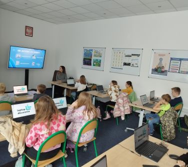 grupa dzieci siedzi przy stolikach w sali lekcyjnej, przy laptopach, na których widnieje napis laptop dla ucznia/zdjęcie...