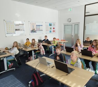 grupa dzieci siedzi przy stolikach z laptopami w sali lekcyjnej/zdjęcie otwiera się w nowej karcie