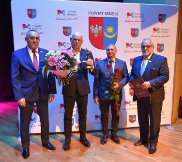 czterech mężczyzn pozuje do zdjęcia dwóch w środku trzyma kwiaty i statuetkę, w tle baner powiat miński