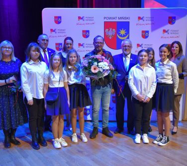 grupa osób pozuje do zdjęcia, mężczyzna w środku trzyma bukiet kwiatów, w tle baner powiat miński