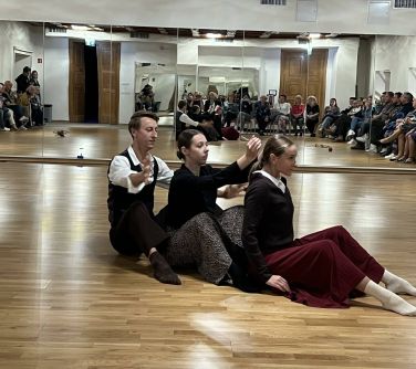 trzy osoby siedzą na podłodze, tańczą, za nimi lustra w ktorych odbija się siedząca publiczność