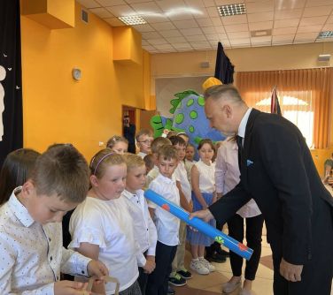 po lewej uczniowie ubrani na galowo, po prawej mężczyzna trzymając długi ołowek dotyka ramienia chłopca