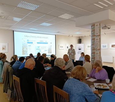 sla konferencyjna, grupa osób siedzi na krzesłach naokoło przy złączonych stołach, z przodu dwie kobiety jedna stoi druga...