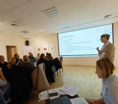 sala konferencyjna, po lewej stronie grupa osób siedzi na krzesłach naokoło stołów, po prawej dwie kobiety, jedna stoi...
