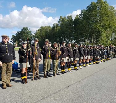 grupa harcerzy stoi w linii na płycie lotniska, w tle drzewa